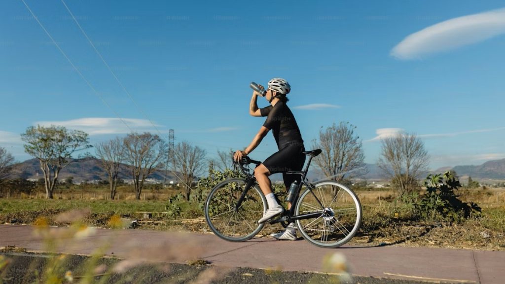 Cycliste en tenue de route noire buvant à son bidon lors d'un entraînement en extérieur pour illustrer l'hydratation et la nutrition sportive.
